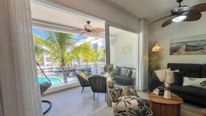 A modern living room with a view of a pool and palm trees, a ceiling fan, and a sofa with decorative pillows. - Dimary Charming Apt, 5 Pools, Walk to the Beach Dominicus Bayahibe la Romana (Dominicus)