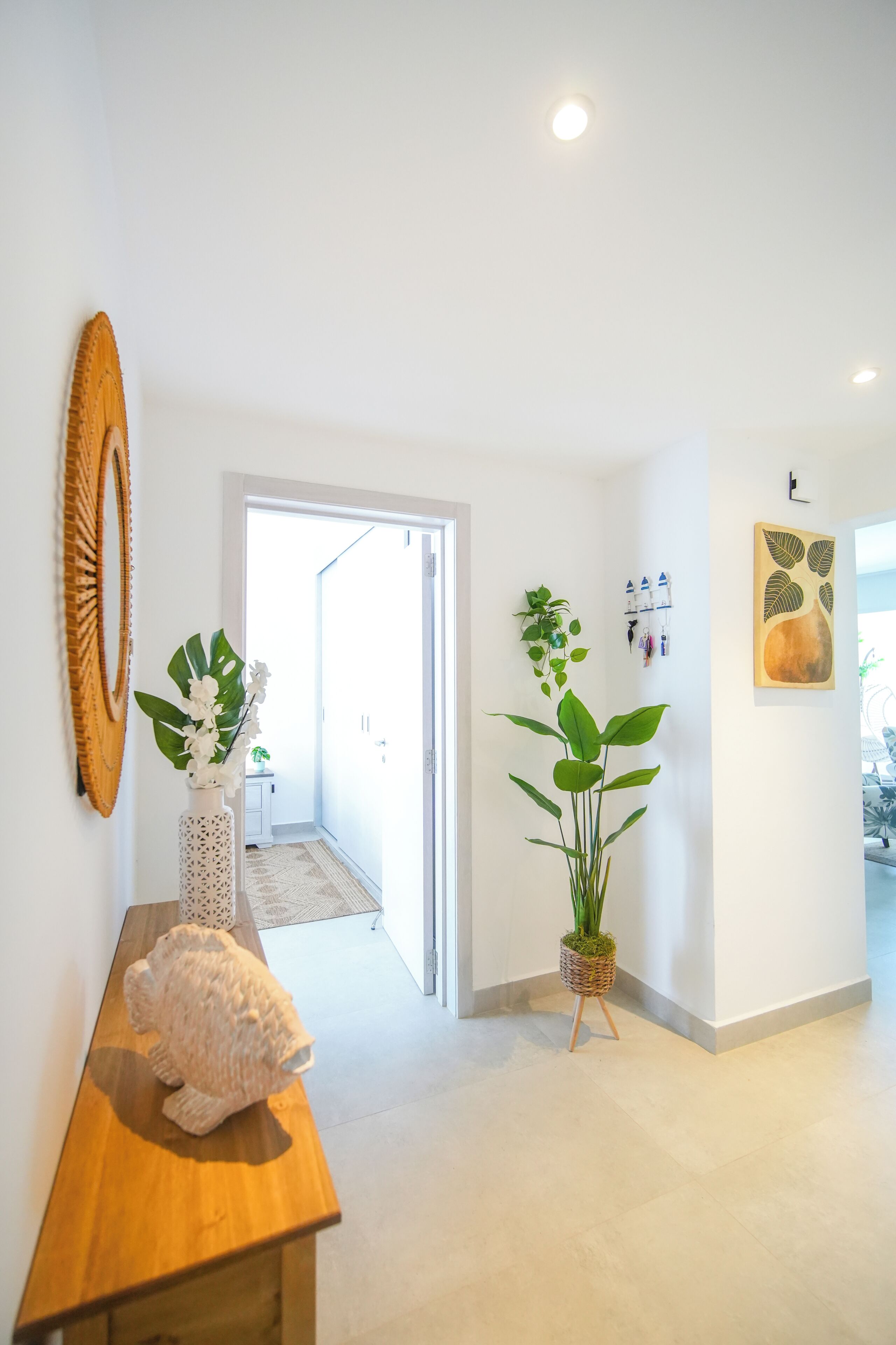 A hallway with a wooden bench, a white vase with orchids, a large potted plant, and a wall-mounted light fixture.