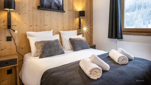 Ein gemütliches Schlafzimmer mit Holzwänden, einem Bett mit weißer und grauer Bettwäsche, zwei Wandlampen und einem Fenster mit Blick auf einen verschneiten Wald.