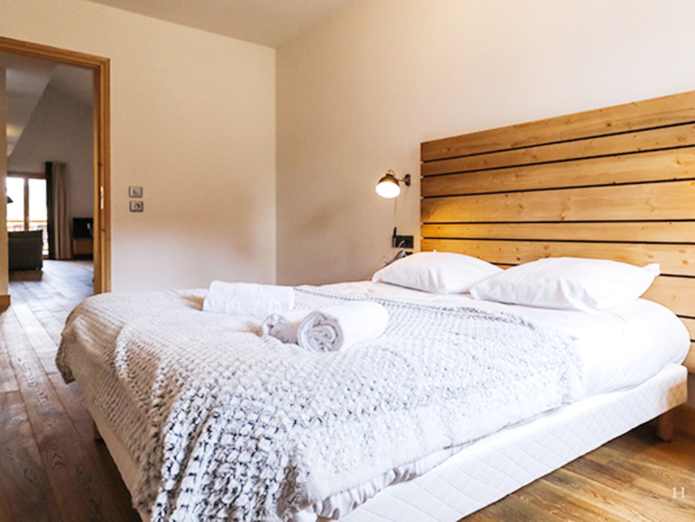 Una cama bien hecha con sábanas blancas y una manta tejida, un cabecero de madera y una lámpara de noche.