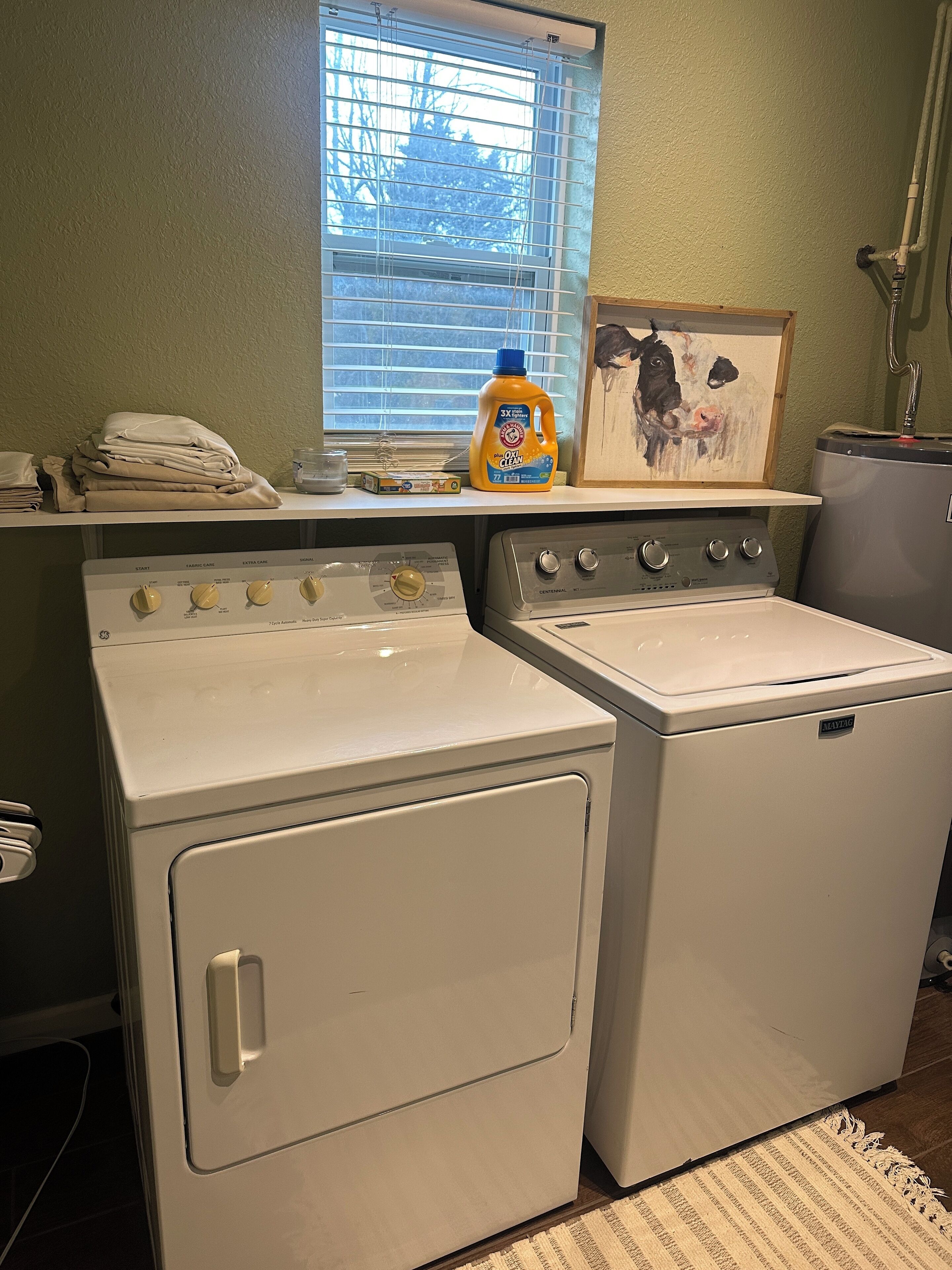 A laundry room with two front-loading washers and dryers, a window with blinds, a shelf with folded clothes, a bottle of detergent, and a framed picture.