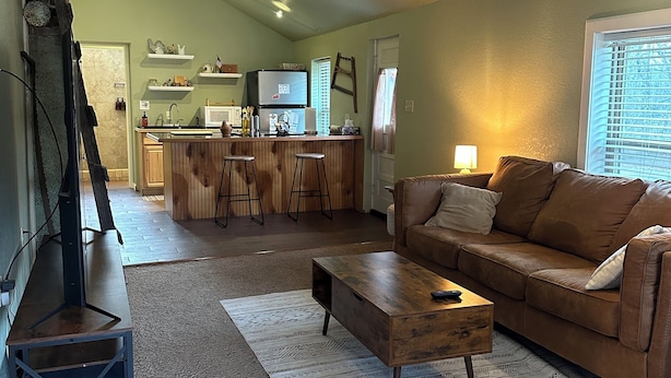 Una sala de estar con un sofá de cuero marrón, una mesa de centro de madera y un mueble para televisor. Se observa una cocina al fondo con un refrigerador, microondas y taburetes de bar.