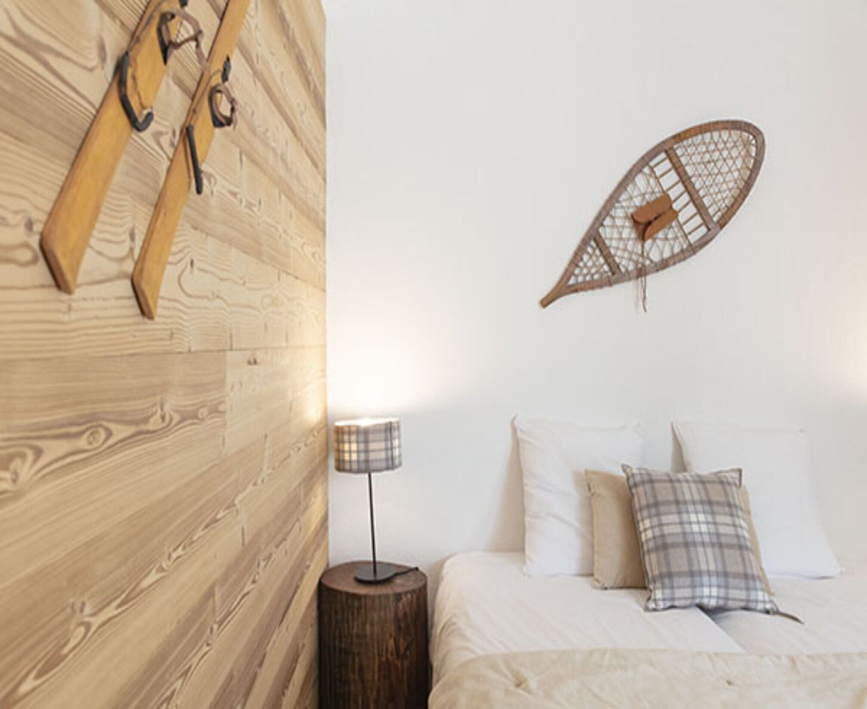 Een slaapkamer met een houten hoofdbord, een bed met wit beddengoed en geruite kussens, een klein nachtkastje met een lamp en een decoratief object aan de muur.