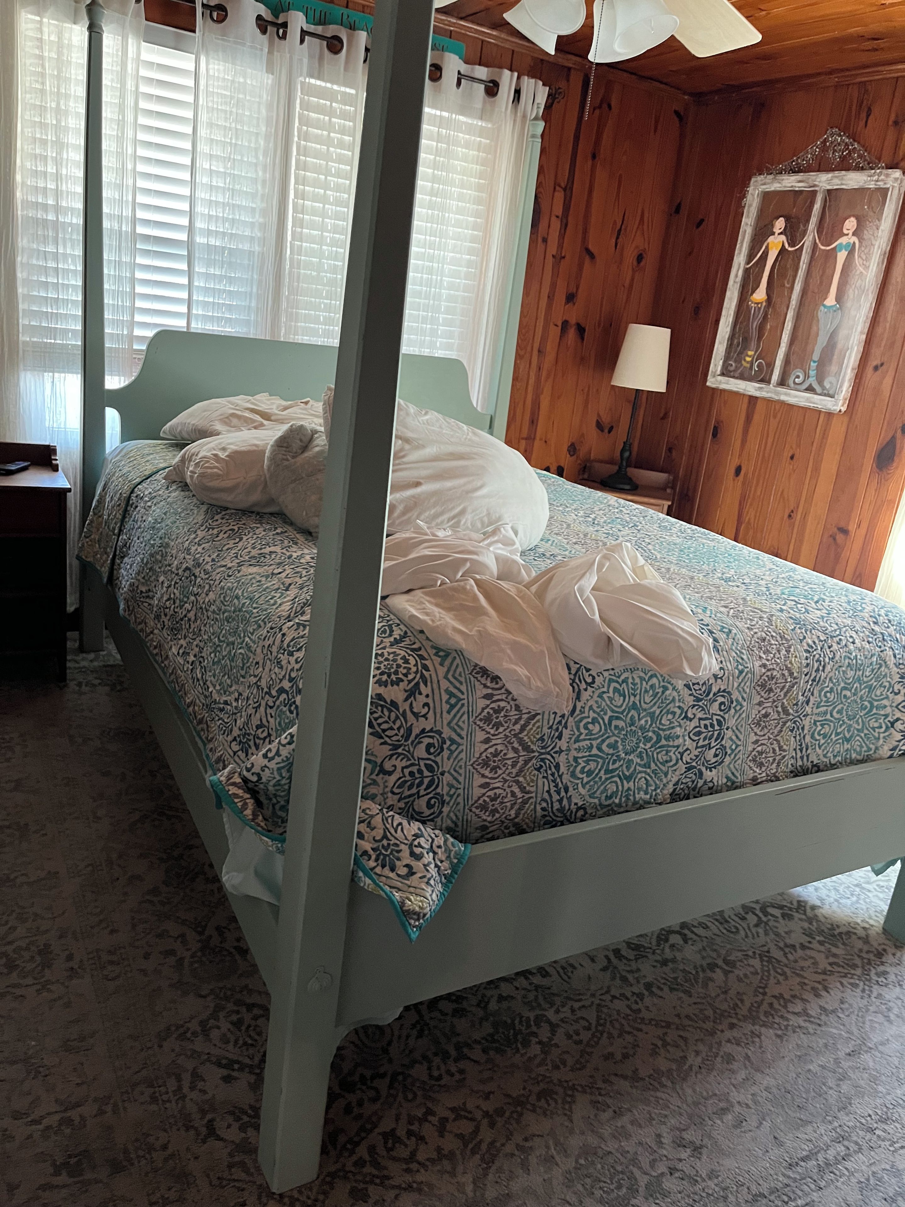 Una cama con dosel con un edredón estampado, una mesita de noche con una lámpara, y una pared de madera con una obra de arte enmarcada.