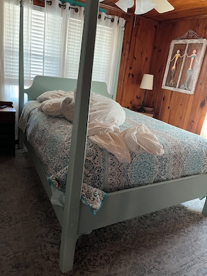 Una cama con dosel con un edredón estampado, una mesita de noche con una lámpara, y una pared de madera con una obra de arte enmarcada.