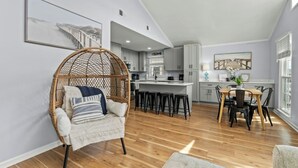 A spacious living area with a wooden floor, a hanging chair, a dining table with chairs, and a kitchen in the background.