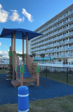 Un parque infantil con un toldo azul y estructuras de juego de madera, un barril azul y un edificio de hotel de varios pisos al fondo.