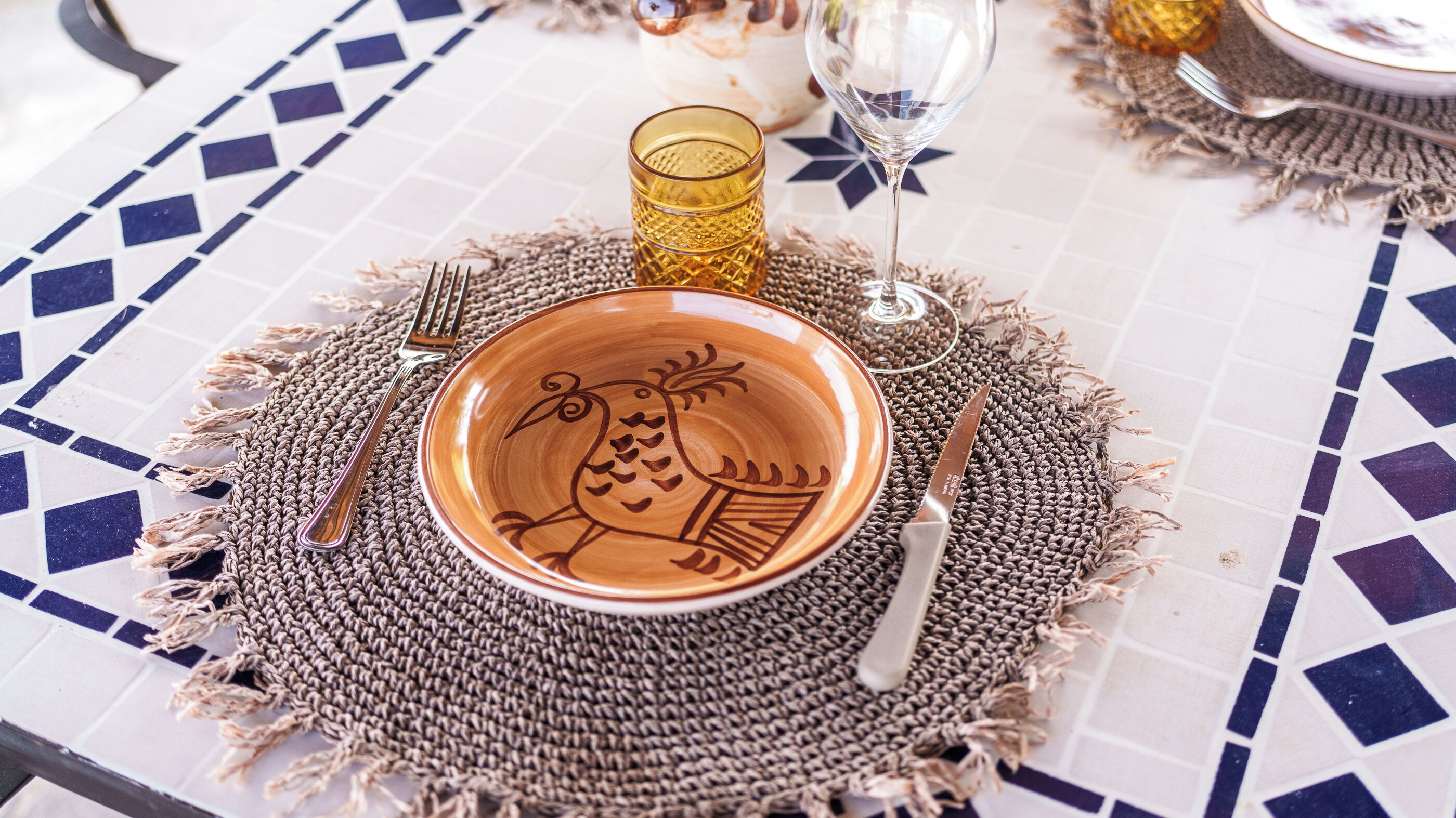 Una mesa con un bol de cerámica, un vaso, un cuchillo y un tenedor sobre un mantel tejido.