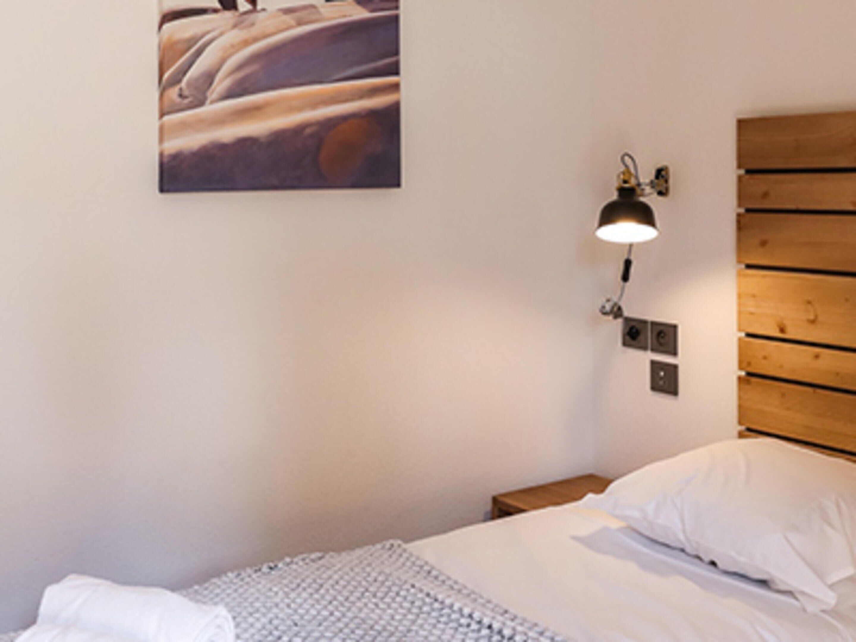 A neatly made bed with white linens, a wooden headboard, a wall-mounted lamp, and a framed picture above the bed.
