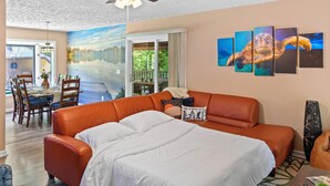 Un salon chaleureux avec un grand canapé orange, un coin repas avec une table et des chaises, et une fresque murale représentant un paysage.