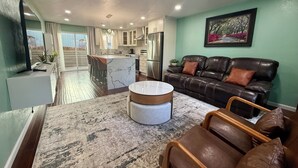 A modern living room with a brown leather sofa, a round ottoman, and a large rug. A kitchen with white cabinets and a marble island is visible in the background.
