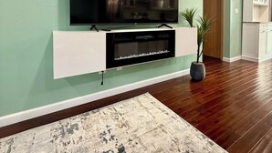 A living room with a flat-screen TV mounted on a white entertainment unit, a patterned rug, and a potted plant.