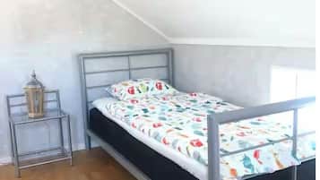 A small, single-bed room with a sloped ceiling, a metal bed frame, a colorful bedspread, a bedside table with a lamp, and a wooden floor.