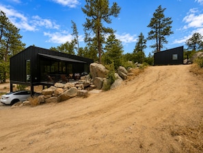 Une cabane moderne dotée d’une terrasse, entourée d’arbres et bordée d’un sentier de terre.