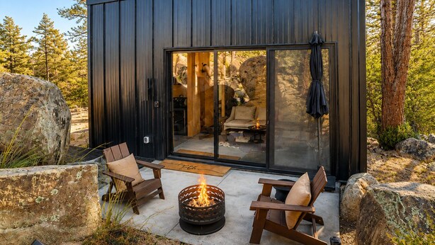 En moderne hytte med bålsted, to træstole og en glasdør ind til et interiør med sofa og spisebord.