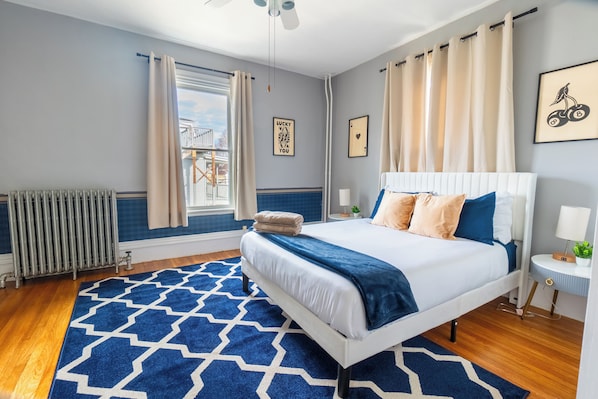 Un dormitorio con una cama grande, un tapete estampado, un radiador, una ventana con cortinas y una pared decorada con cuadros.