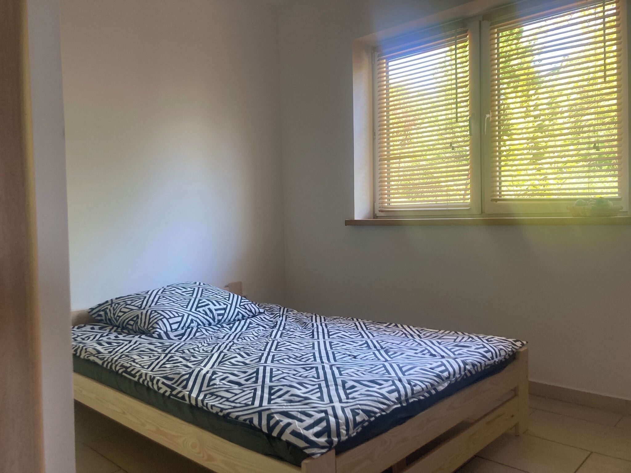 A single bed with a geometric patterned bedspread in a room with white walls and a window with horizontal blinds.