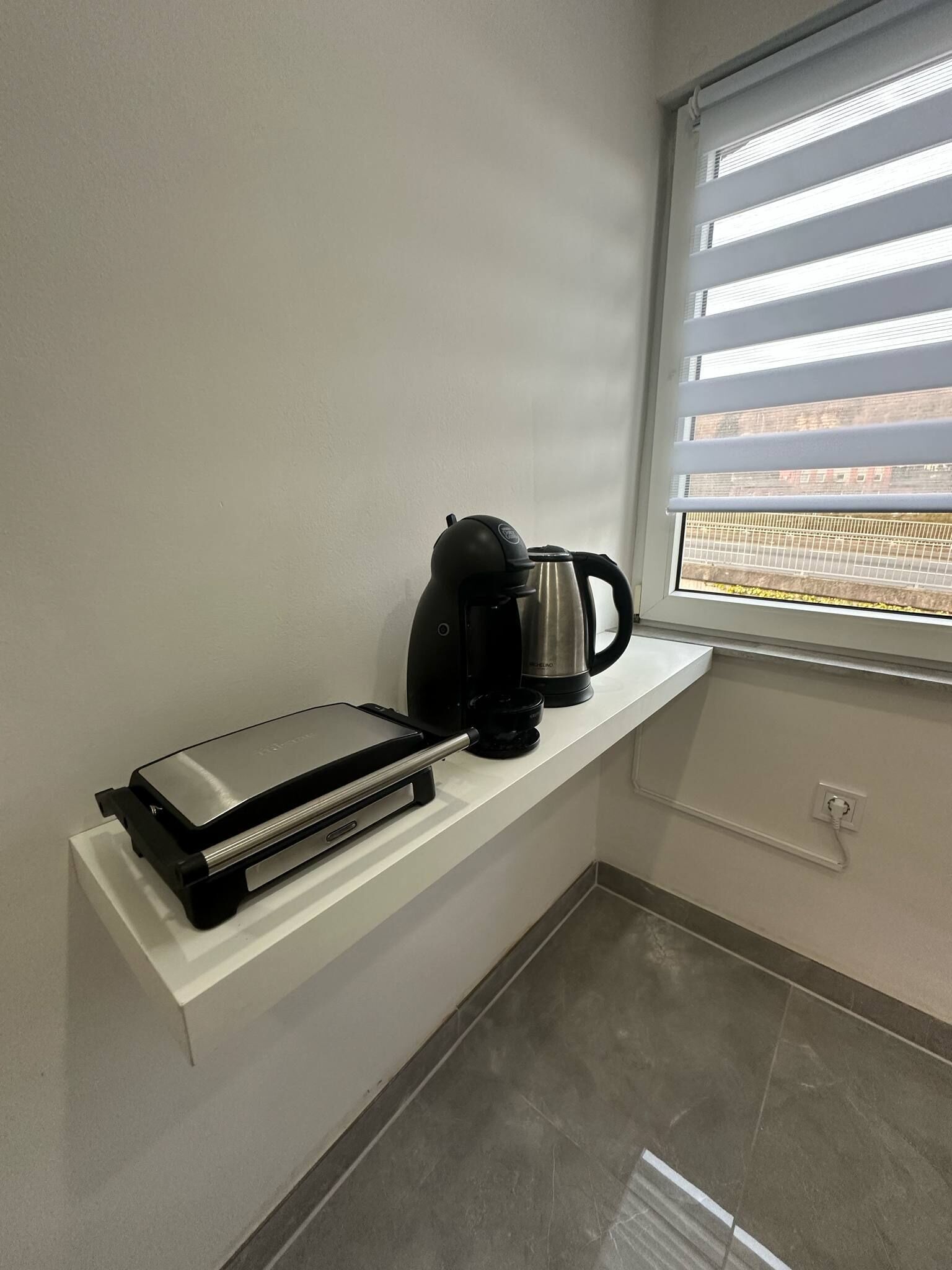 Una kitchenette con cafetera, hervidor y tostadora sobre una encimera blanca, junto a una ventana con persianas.