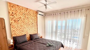 A bedroom with a wooden headboard, a bed with dark bedding, a ceiling fan, and a view of the city through sheer curtains. - Vista Adriatic CZ (Durres)