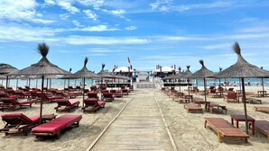 A beach with wooden lounge chairs and thatched umbrellas, a wooden walkway leading to a building, and a clear blue sky. - Vista Adriatic CZ (Durres)
