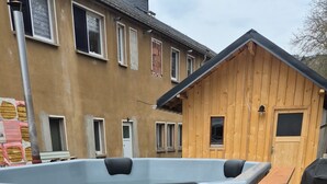 Una bañera de madera sobre un remolque con estructura metálica, una terraza de madera y una casa al fondo.