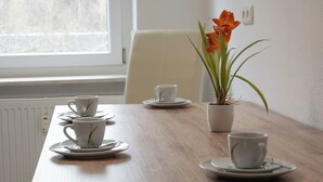 Una mesa de comedor con tazas y platillos blancos, una planta en maceta con flores naranjas y una silla blanca.