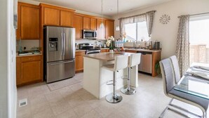 A modern kitchen with wooden cabinets, stainless steel appliances, a central island with bar stools, and a dining area with a glass table. - Wake up to the best panoramic views in this cozy 3-bedroom house. (West Jordan)