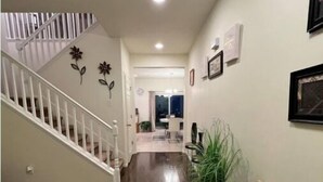 A hallway with a wooden floor, a staircase with white railings, a beige sofa with decorative pillows, and a plant. - Wake up to the best panoramic views in this cozy 3-bedroom house. (West Jordan)
