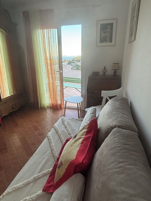 A living room with a sofa, a chair, a small table, and a view of a balcony with a green bench.