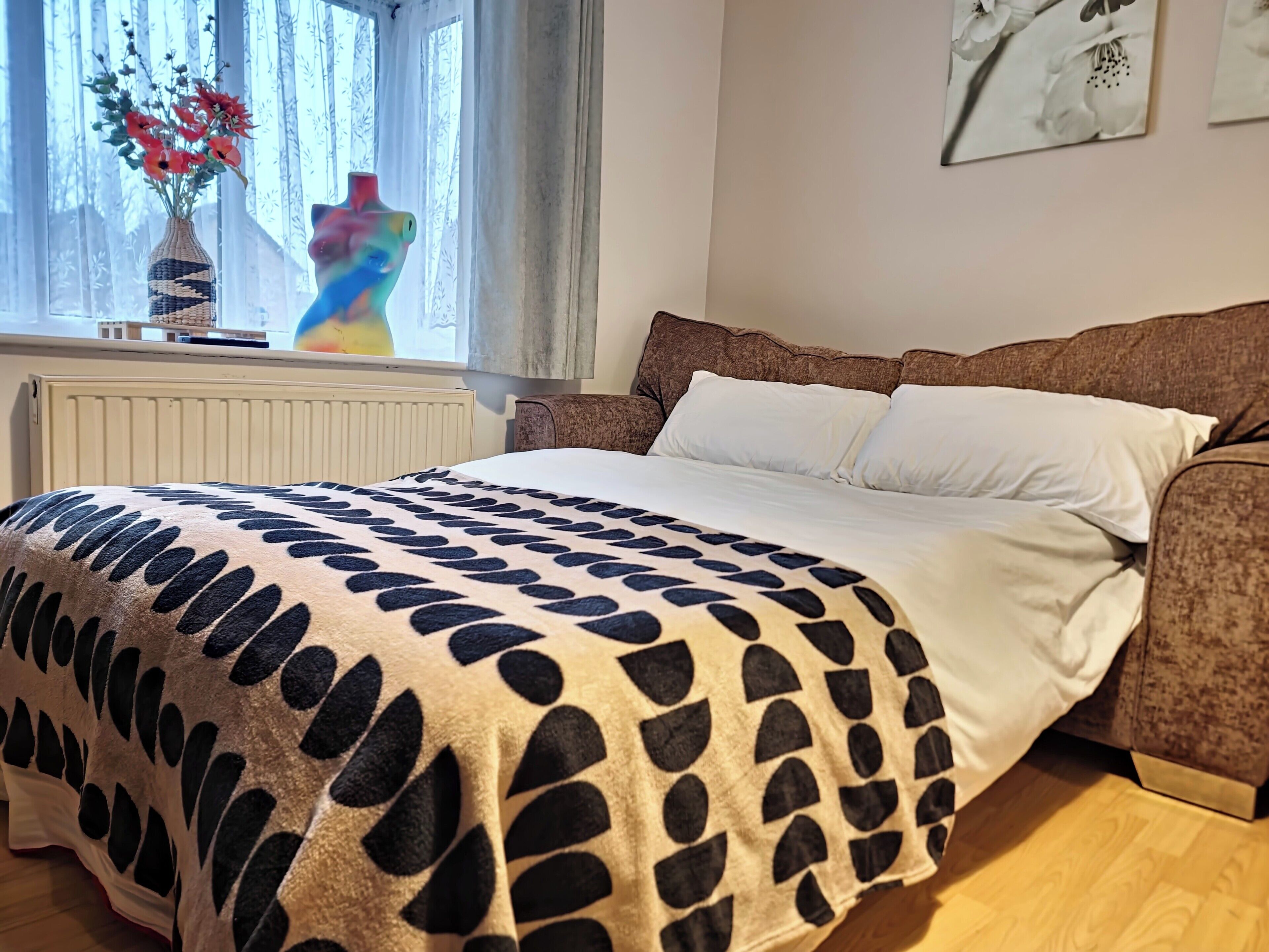 Une chambre à coucher avec un lit, une fenêtre avec des rideaux, un radiateur et un mur orné d’un tableau encadré.