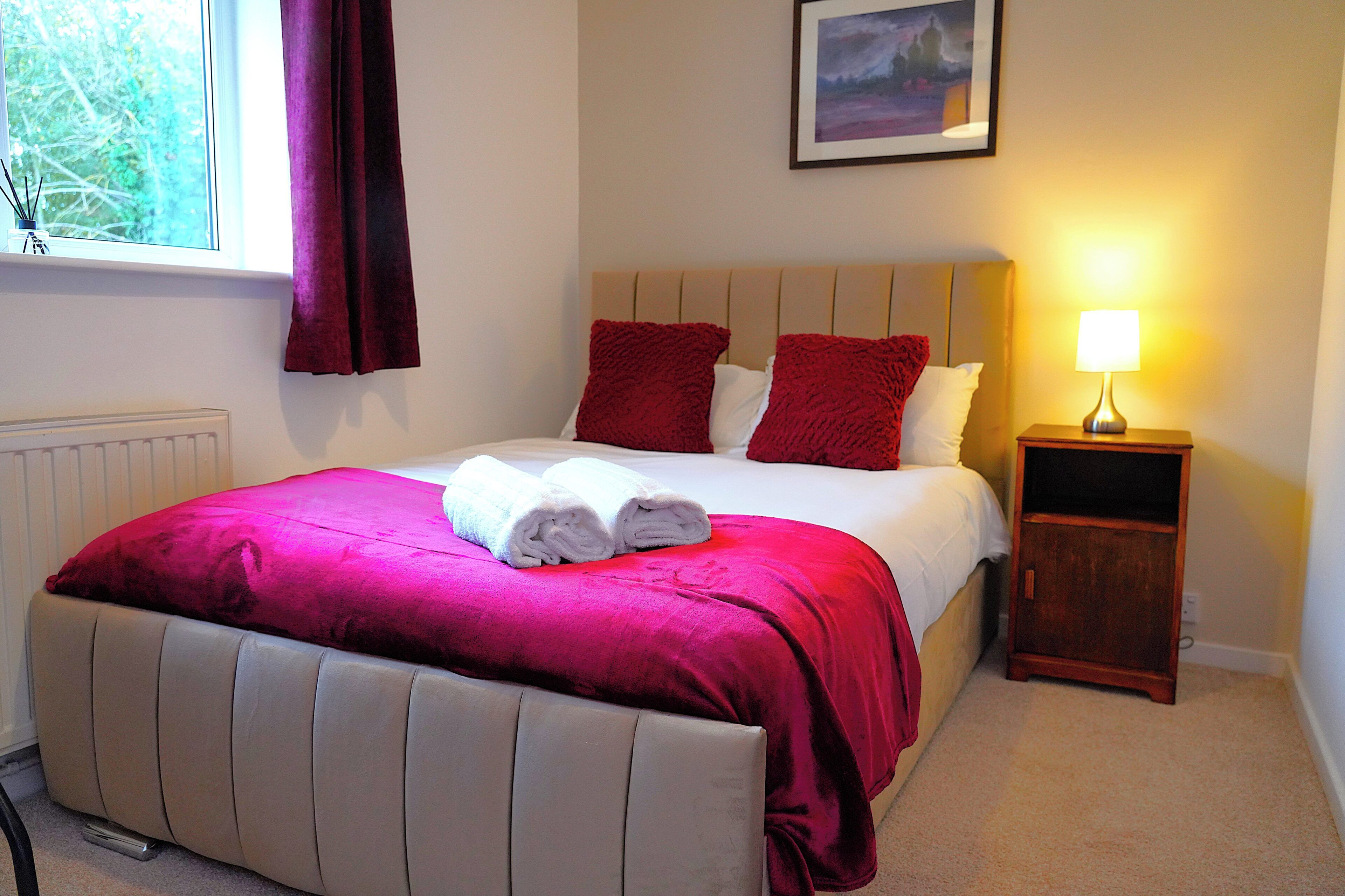 A neatly made bed with a red and white bedspread, a wooden nightstand with a lamp, and a framed picture on the wall.