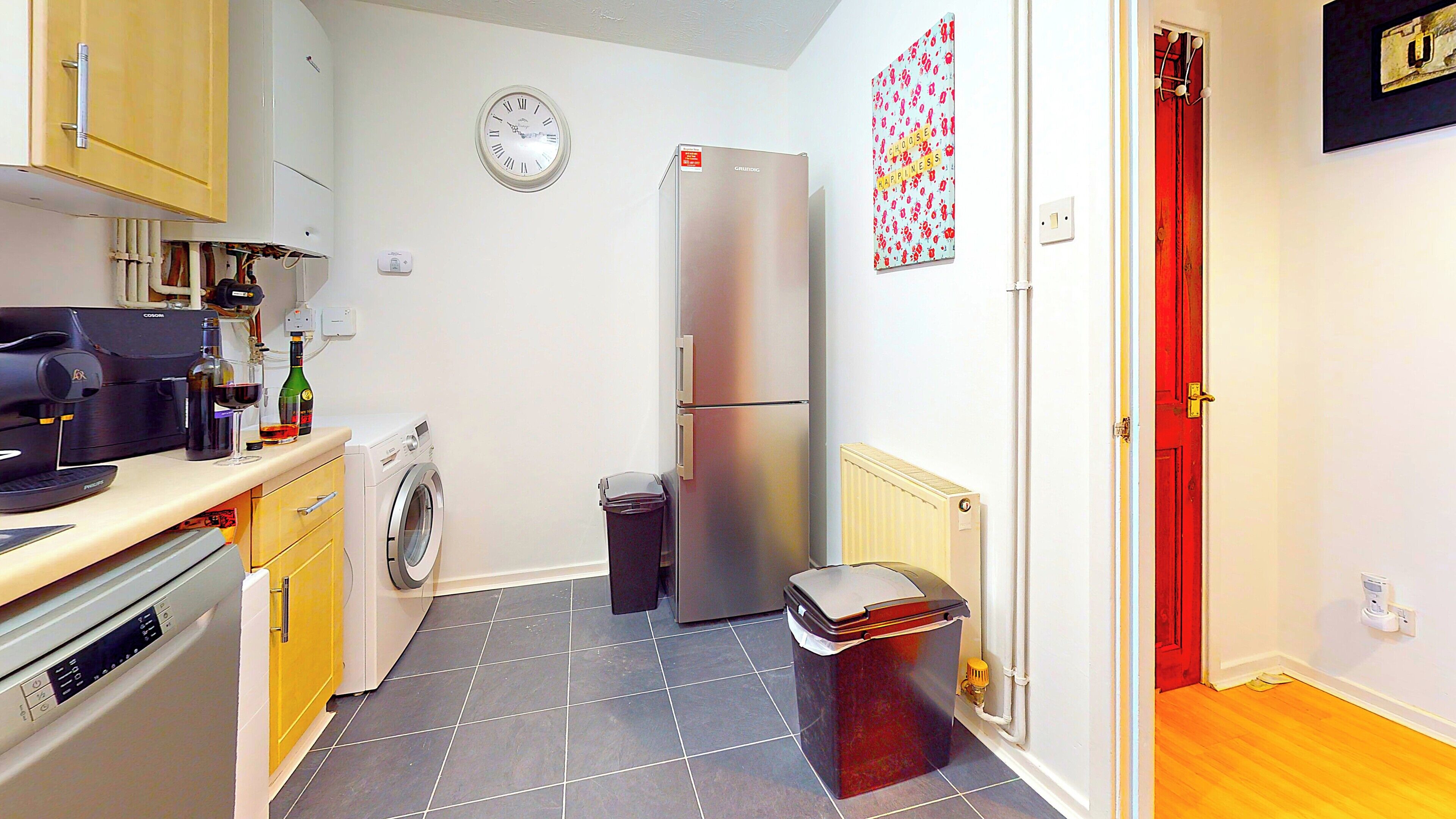 A compact kitchen with a washing machine, a refrigerator, a coffee machine, and a clock on the wall.
