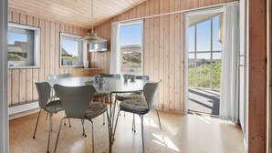 Un espace de repas moderne avec une table ronde et six chaises, de grandes fenêtres offrant une vue sur la verdure et un plafond en bois.