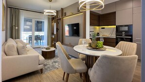 Een moderne woonkamer met een bank, stoelen, een eettafel met fruit, een televisie en een keuken met apparatuur.