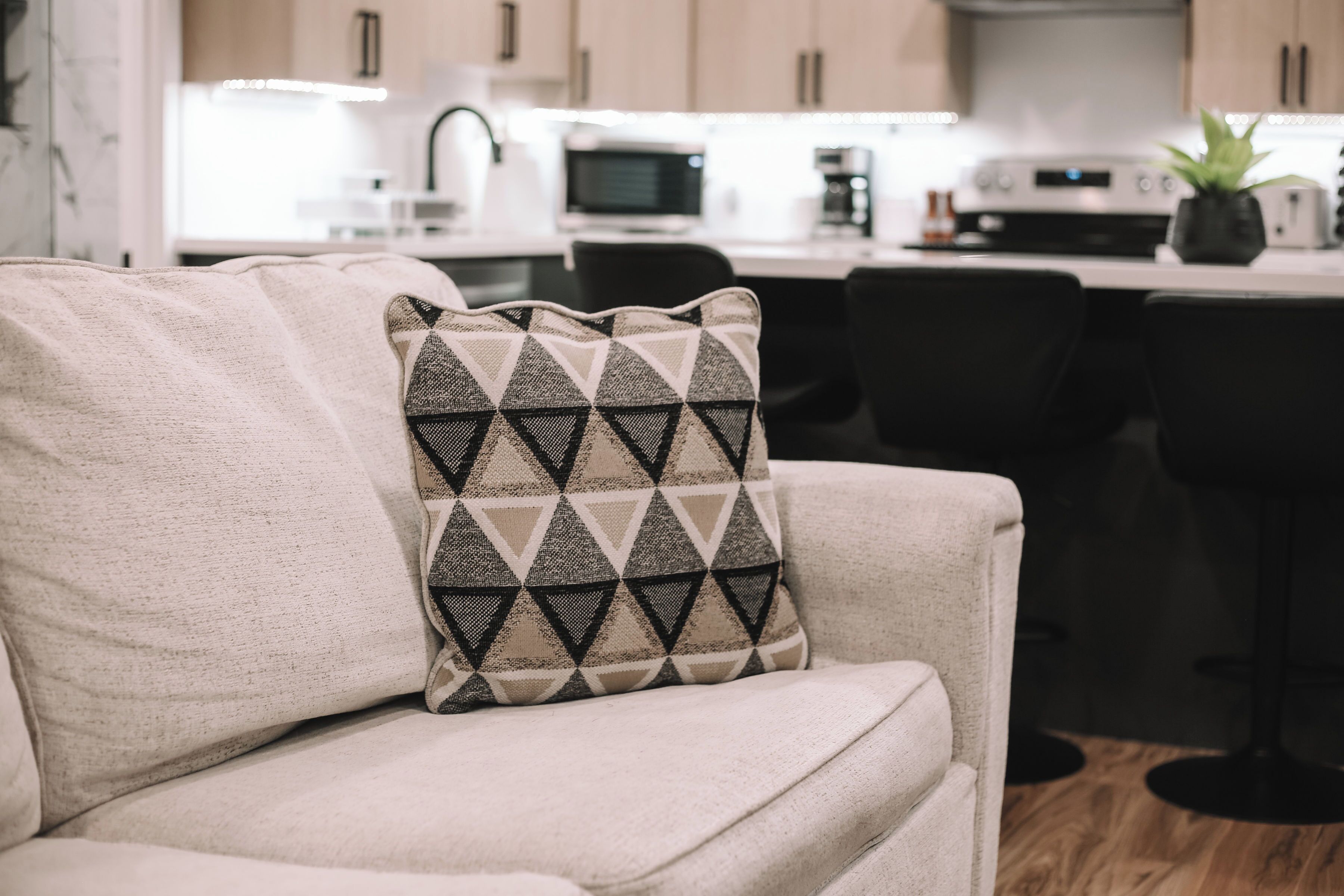 A living room with a sofa and a patterned pillow, a kitchen in the background with modern appliances.