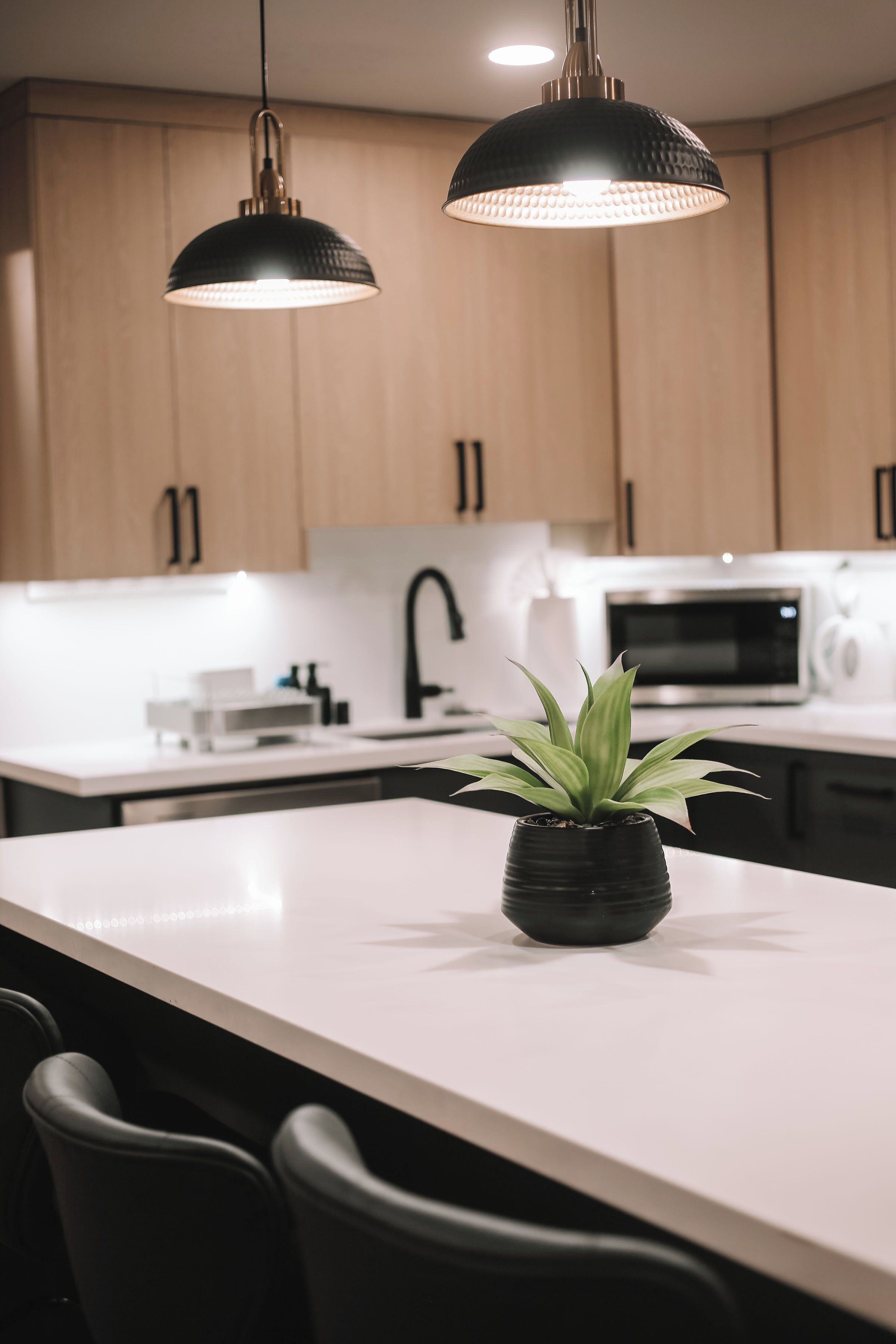 Une cuisine moderne avec des meubles en bois, un plan de travail blanc, deux lampes suspendues, une plante en pot et un four à micro-ondes.