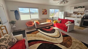 A living room with a sofa, armchairs, a coffee table, and a rug. There are windows, a ceiling fan, and a kitchen area visible. - Mountain View Retreat Near Zion, Sand Hollow (Hurricane)