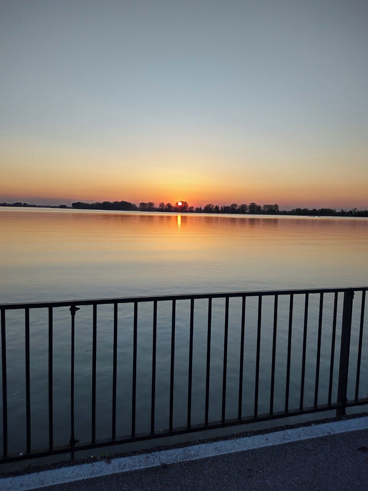 Una masa de agua tranquila con un atardecer al fondo y una baranda de metal en primer plano.