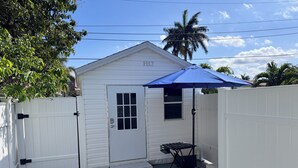 A small white building with a door and a window, a blue umbrella, and a black metal table.