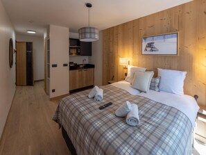 A hotel room with a wooden wall, a bed with a plaid bedspread, two rolled towels, a remote control, a painting of a car on the wall, and a kitchenette in the background. - Apartment in Serre-chevalier Near Ski Slopes (Saint-Chaffrey)