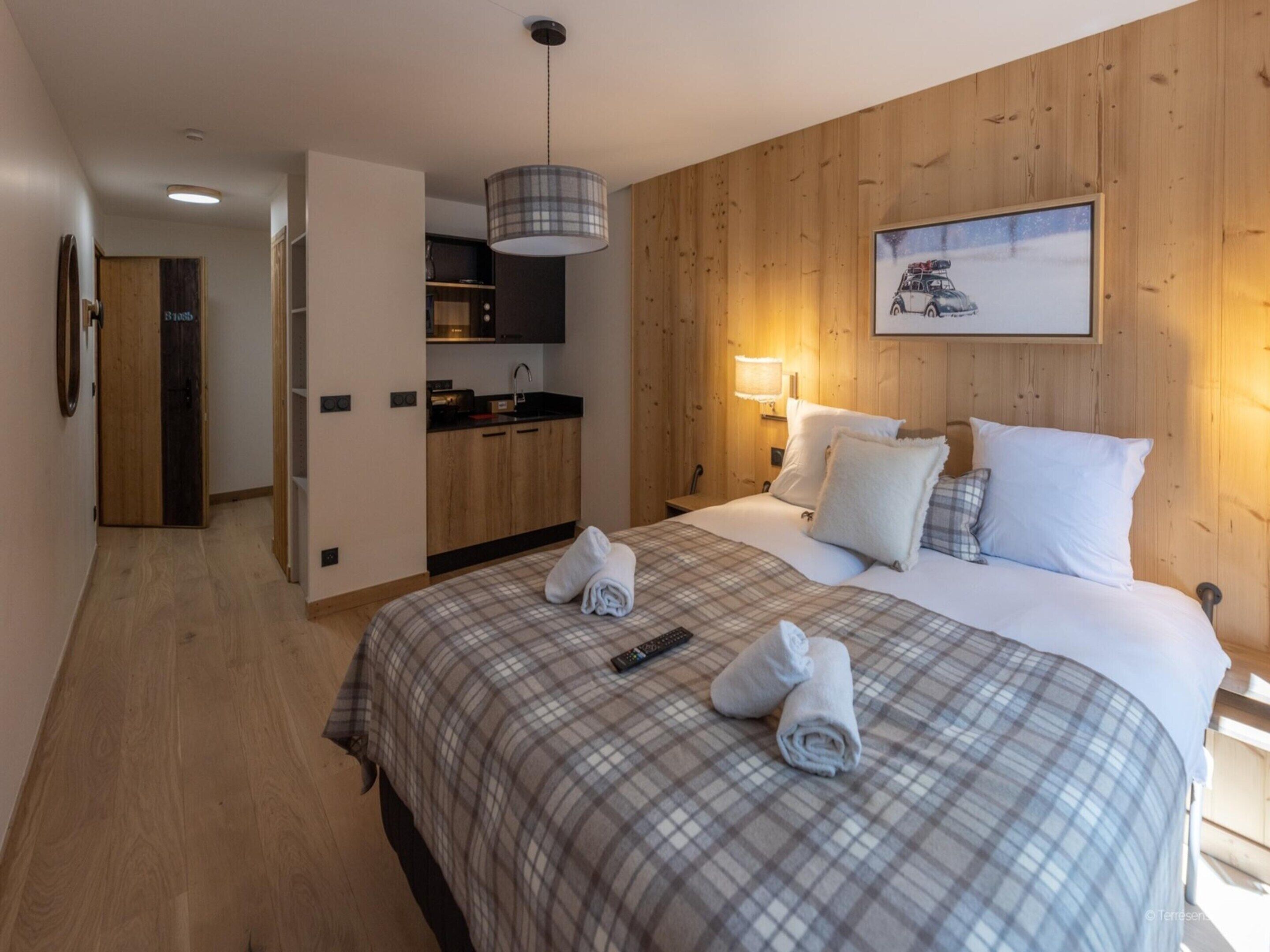 A hotel room with a wooden wall, a bed with a plaid bedspread, a painting, a kitchen area, and a hallway.