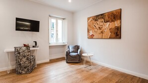 A modern living room with hardwood flooring, a flat-screen TV, a window, a chair, a small round table, and a large abstract painting. - Jacuzzi Suite – Nikis Collection Trastevere (Roma)