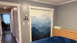 Un dormitorio con cama, cómoda, puerta con un mural de montaña y un pasillo que conduce a otra habitación.