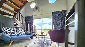 A modern living room with a wooden staircase, a blue sofa with patterned cushions, a purple chair, and a large window offering a view of the outdoors. - Charming 1-bedroom chalet in lush Poas with cascading pools & raw nature  WiFi (Poas)
