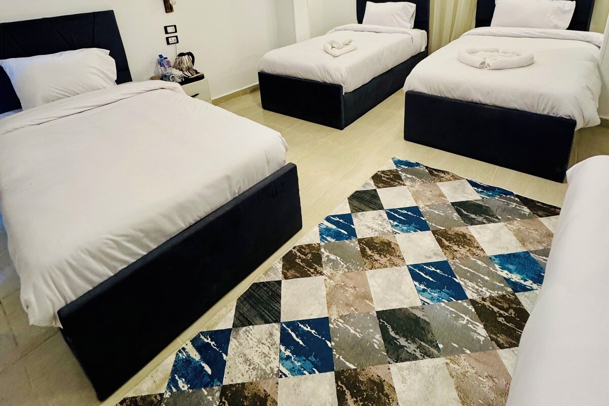 A hotel room with two beds, a large window with curtains, a patterned rug, and a wall-mounted air conditioning unit.