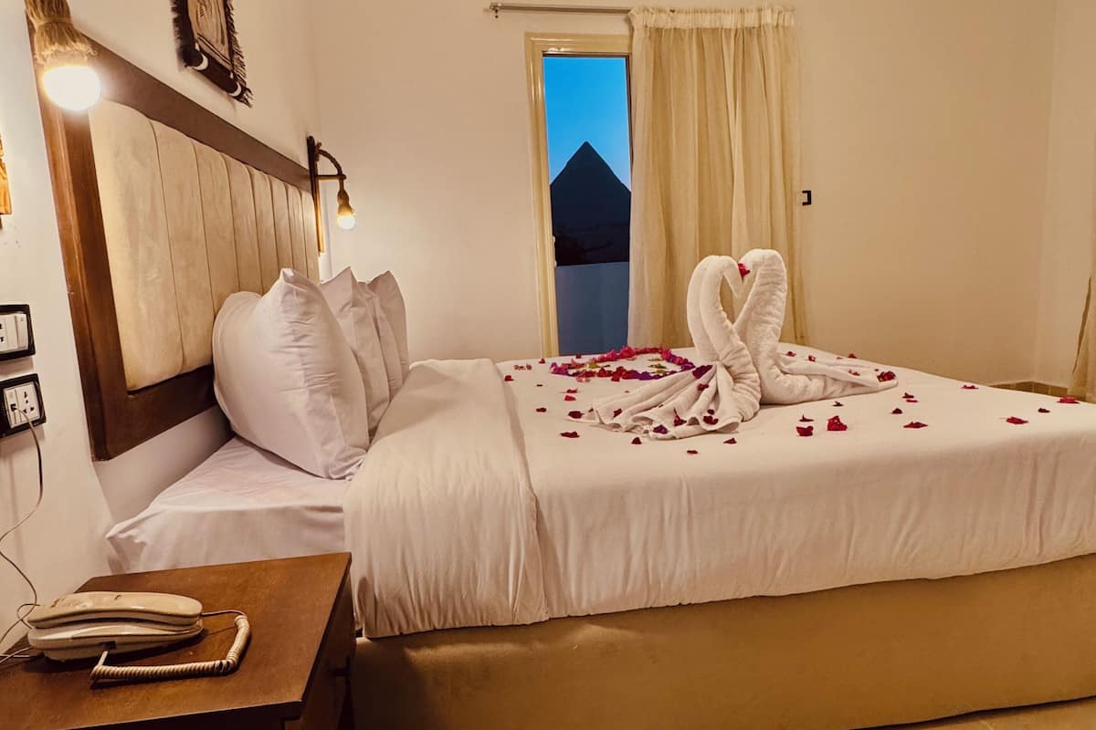 A hotel room with a bed, a nightstand, a telephone, and a view of a pyramid through the window.