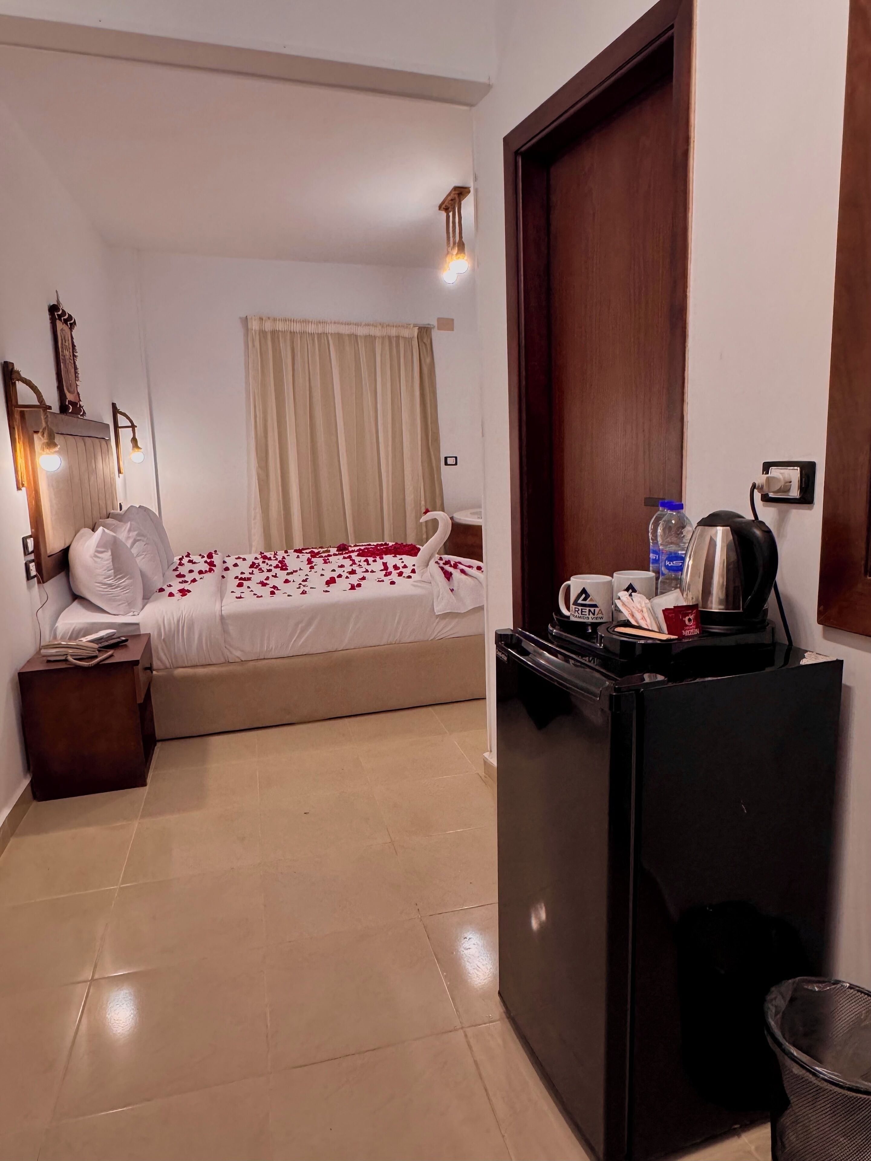 A hotel room with a bed, a small table with a kettle, a trash bin, and a door.