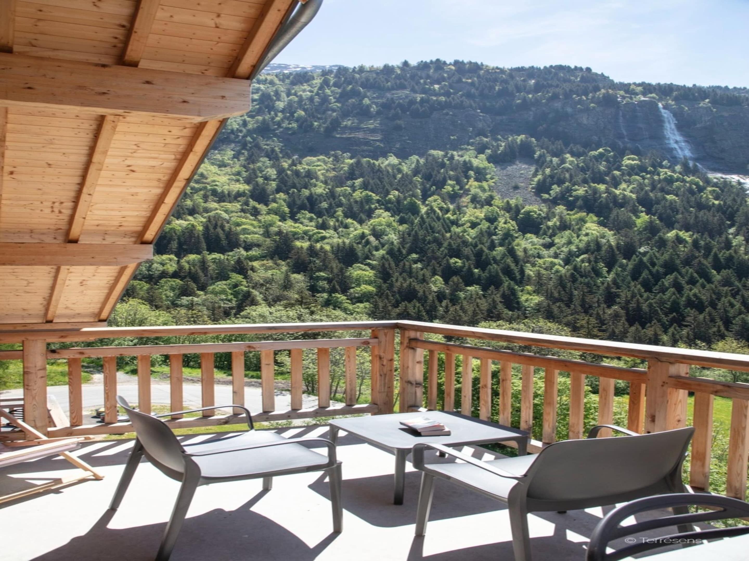 Eine Holzterrasse mit Gartenmöbeln, mit Blick auf eine bewaldete Berglandschaft und einen Wasserfall.