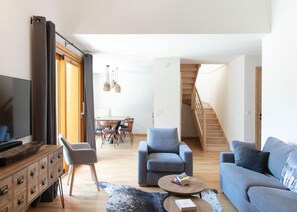 A modern living room with a sofa, armchairs, a coffee table, and a TV. There's a dining area with a table and chairs, and a staircase leading upstairs. - Apartment in Vaujany Near Alpe D'huez Ski Area (Vaujany)