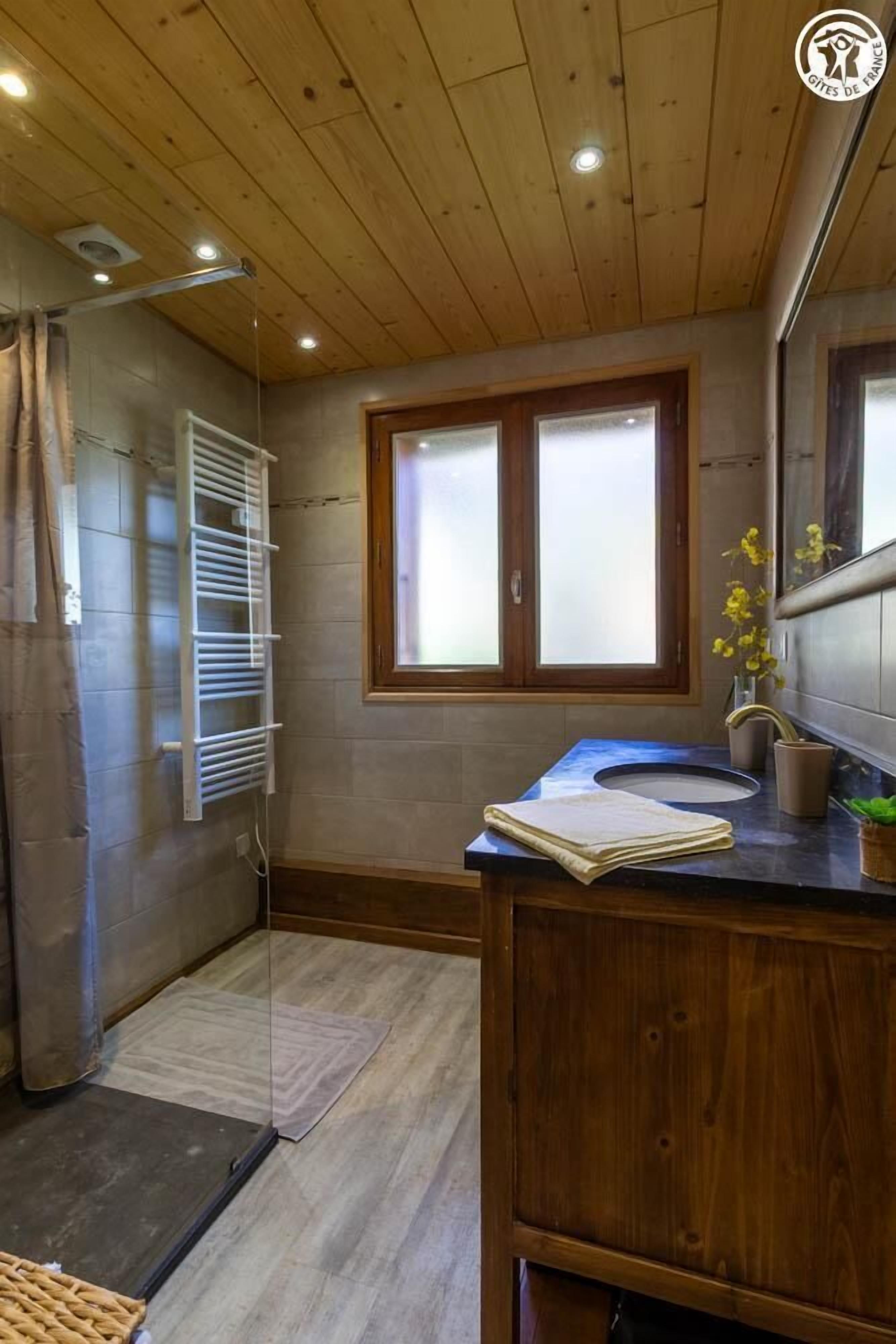 A bathroom with a wooden ceiling, a window with frosted glass, a towel rack, a sink with a mirror, and a small plant.